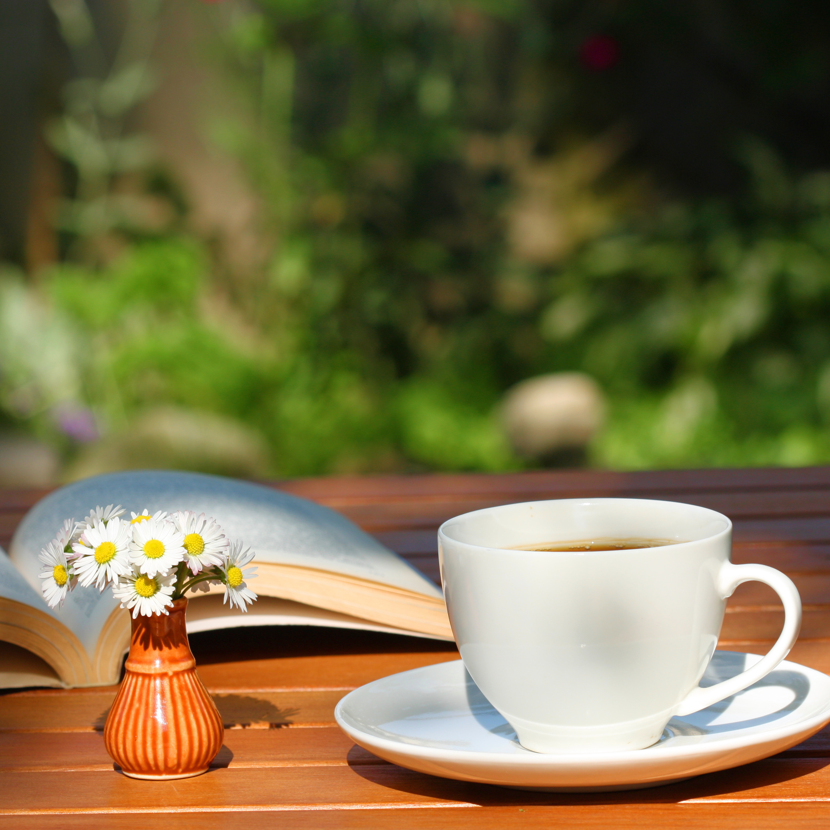A cup of coffee, an open book and a small vase of daisies on a garden table, illustrating taking time to relax in a summer garden.