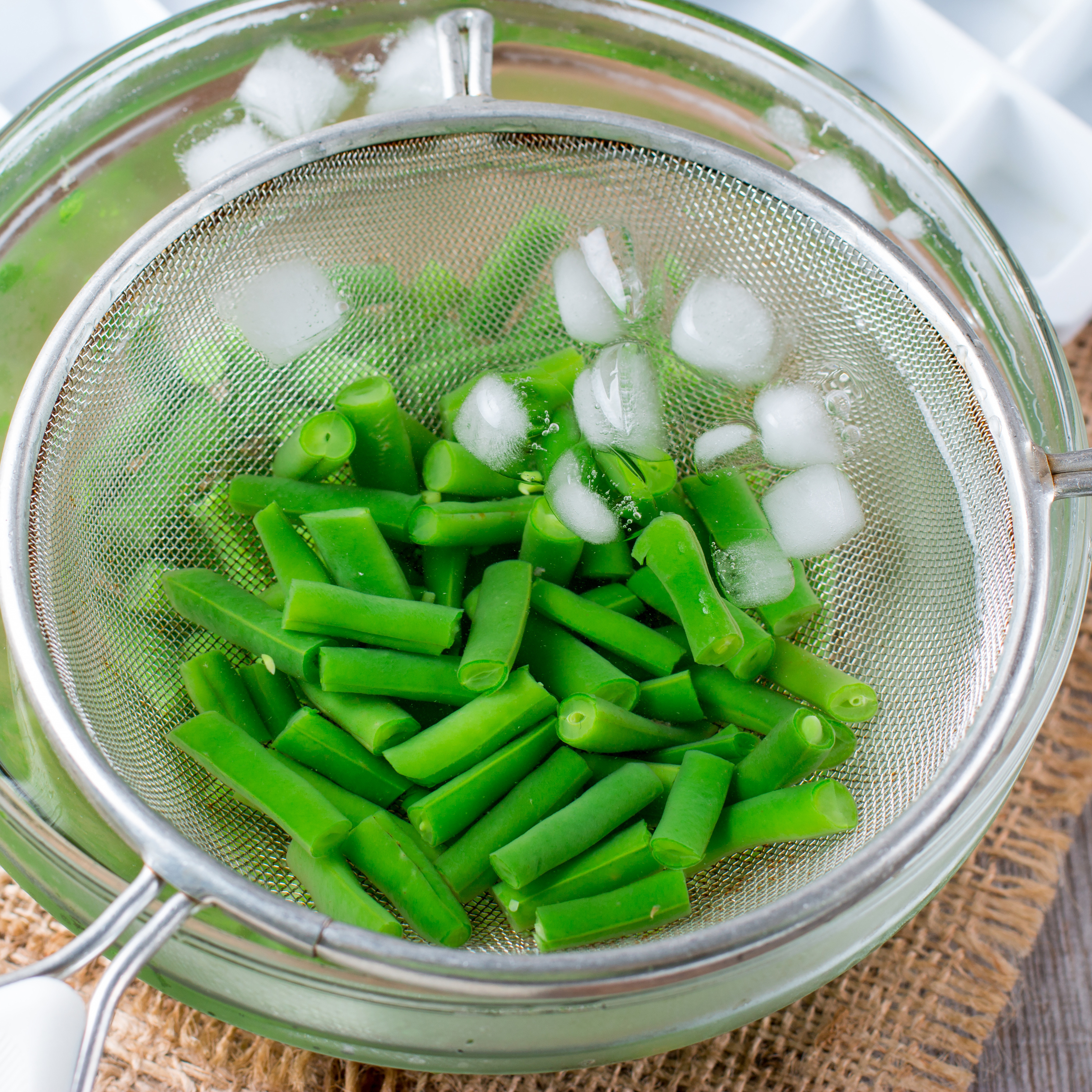 Blanched green beans cooling in an ice-water bath with a mesh strainer.